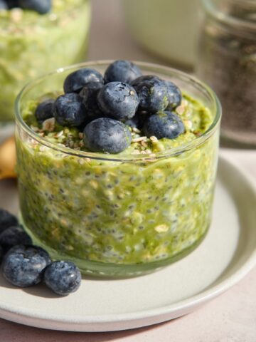 A clear glass jar filled with vibrant green matcha overnight oats speckled with chia seeds and rolled oats, topped with a generous mound of fresh plump blueberries and a sprinkle of seeds, placed on a sage green ceramic plate with additional blueberries alongside and a gold spoon, with a second jar of matcha oats and a glass jar of chia seeds softly blurred in the background on a light pink surface.