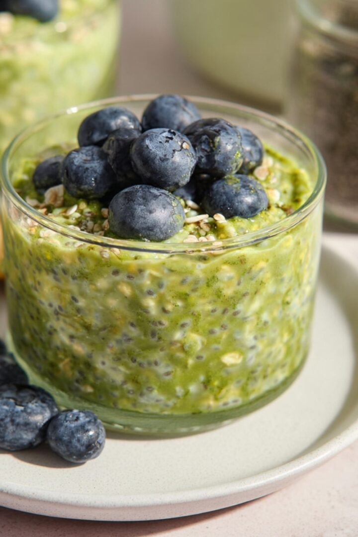 A clear glass jar filled with vibrant green matcha overnight oats speckled with chia seeds and rolled oats, topped with a generous mound of fresh plump blueberries and a sprinkle of seeds, placed on a sage green ceramic plate with additional blueberries alongside and a gold spoon, with a second jar of matcha oats and a glass jar of chia seeds softly blurred in the background on a light pink surface.