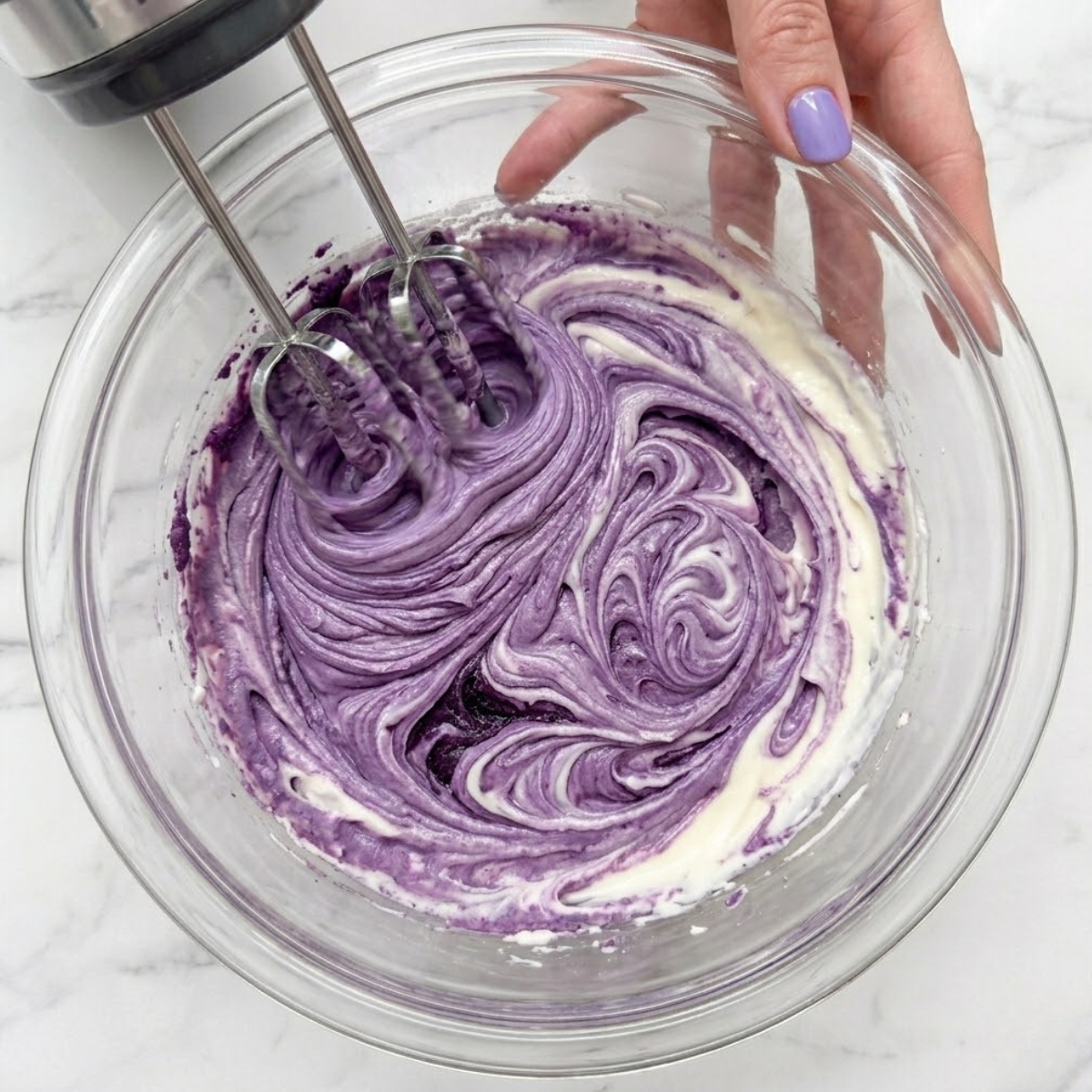 Extreme close-up of hands with purple nails using a hand mixer to blend deep purple ube jam into cream cheese batter.