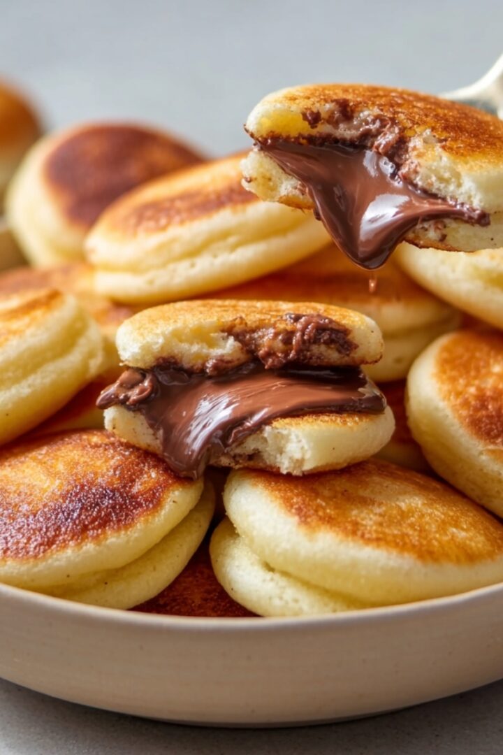 A bowl of golden Nutella Filled Mini Pancake Bombs, with one split open revealing a gooey Nutella center, and a spoon lifting another dripping with melted chocolate.