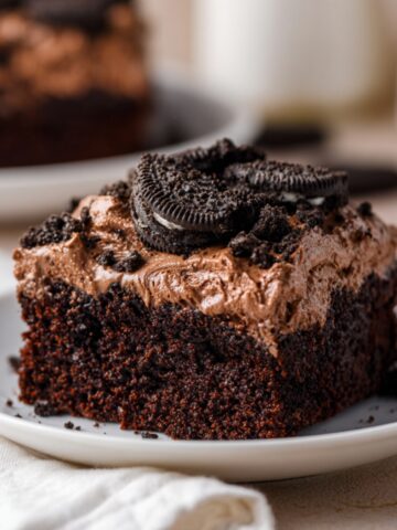 A generous square slice of deep dark chocolate Oreo dirt cake on a white ceramic plate, featuring a dense moist chocolate cake base topped with swirled chocolate cream frosting sprinkled with crushed Oreo crumbs and garnished with a whole Oreo cookie on top, with the remaining cake and a glass of milk softly blurred in the background on a cream linen surface.