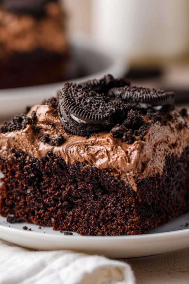 A generous square slice of deep dark chocolate Oreo dirt cake on a white ceramic plate, featuring a dense moist chocolate cake base topped with swirled chocolate cream frosting sprinkled with crushed Oreo crumbs and garnished with a whole Oreo cookie on top, with the remaining cake and a glass of milk softly blurred in the background on a cream linen surface.