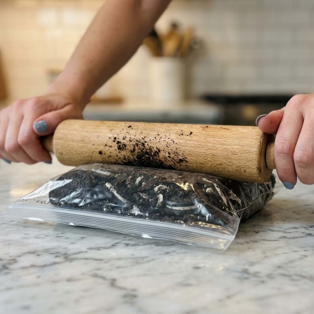 Close-up of hands with blue nails using a rolling pin to crush Oreo cookies in a plastic bag on a marble countertop.