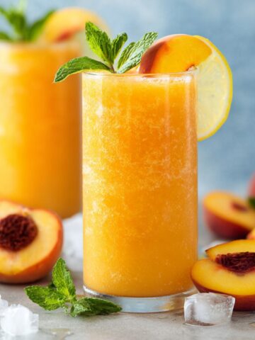 Two tall glasses of frozen Peach Lemonade with a golden orange hue, garnished with a fresh peach slice, lemon wheel, and mint, surrounded by fresh peaches and ice cubes.