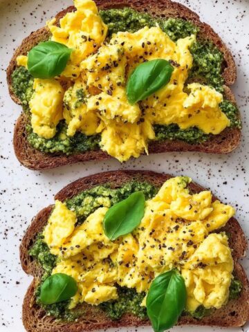 Overhead view of two slices of dark whole grain toast on a white speckled ceramic plate, each spread with a layer of vibrant green basil pesto and topped with fluffy bright yellow scrambled eggs seasoned with cracked black pepper, garnished with fresh whole basil leaves.