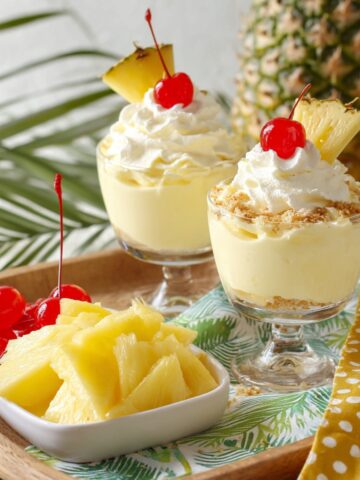 Two glass dessert cups filled with creamy pina colada cheesecake mousse layered over graham cracker crumble, topped with whipped cream, maraschino cherries, and pineapple wedges, served alongside a bowl of pineapple chunks with a tropical pineapple and palm leaf backdrop.