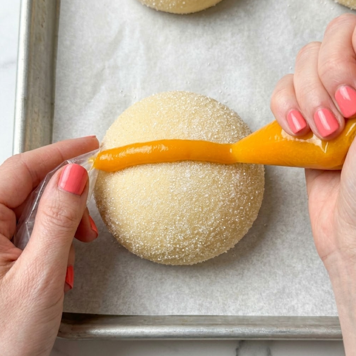Tight zoom of hands with pink nails piping a mango swirl onto a sugar-rolled cookie ball.