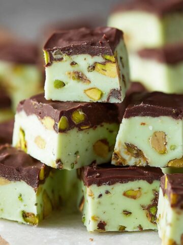 A stacked pile of pistachio fudge squares featuring a pale sage green pistachio-studded fudge base topped with a smooth dark chocolate layer, with chopped green and golden pistachio pieces visible throughout the creamy interior, arranged on white parchment paper on a wooden board with additional pieces softly blurred in the gray background.You said: Pistachio Fudge