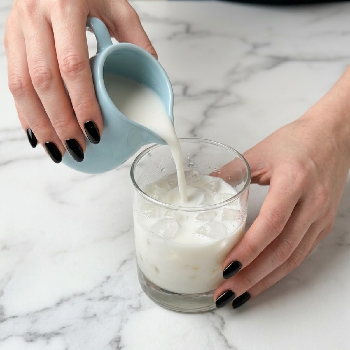 Close up view of a woman's hands with glossy black nails pouring a creamy white coconut milk blend into a clear tumbler glass filled with ice cubes.
