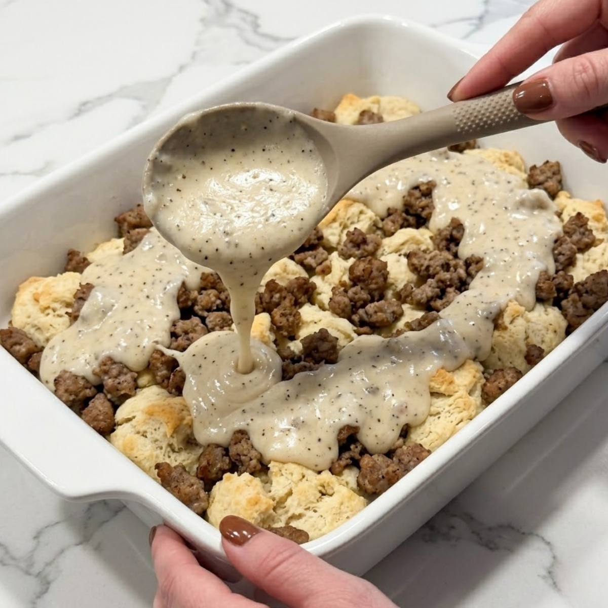 Tight zoom of hands with brown nails pouring thick white country gravy over sausage and biscuit pieces in a white baking dish.