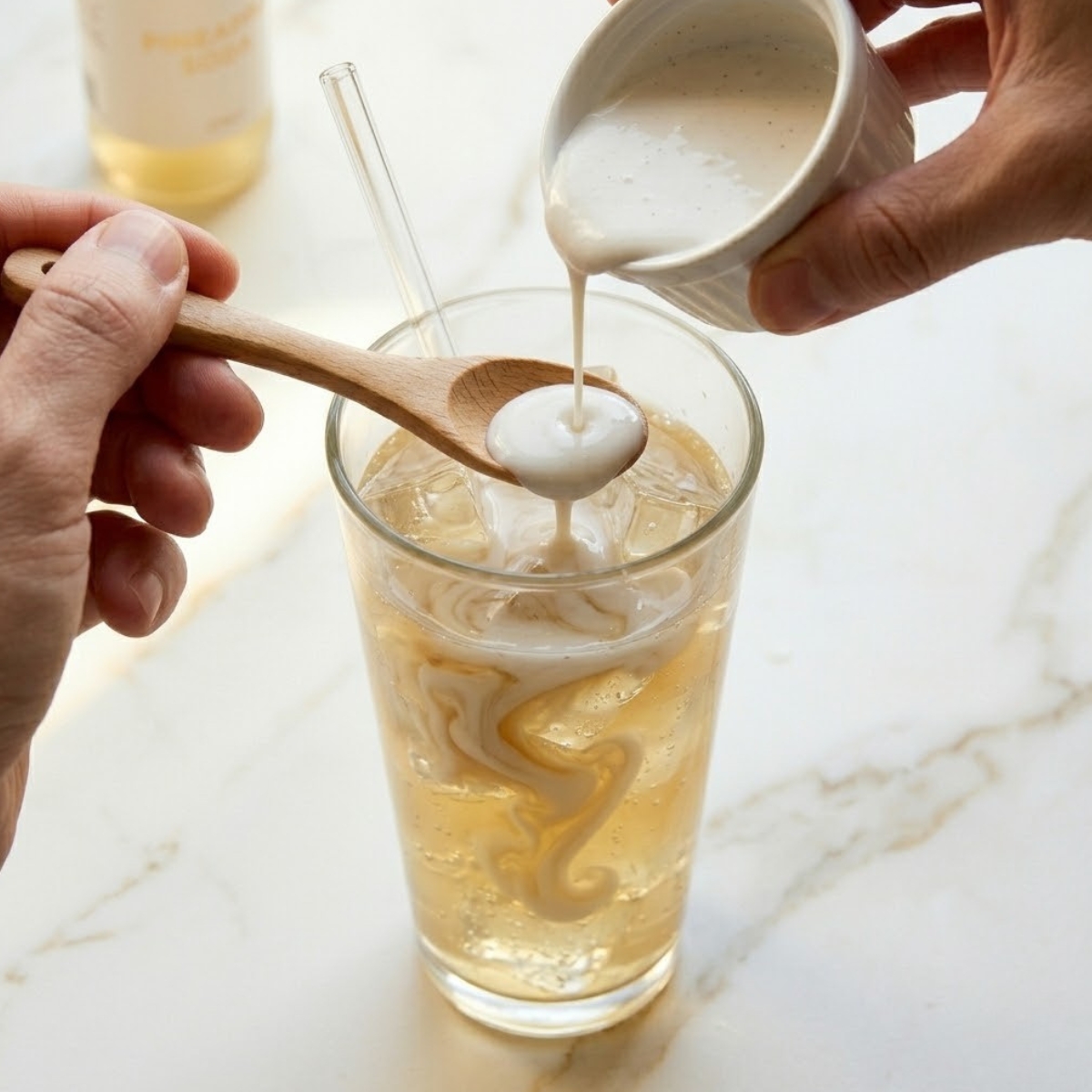 A close-up shot of white coconut cream being poured from a spoon held just above the surface of the golden pineapple soda in a tall glass. Natural daylight from image_10.png illuminates the cream sinking in long ribbons, creating a beautiful marble pattern against the ice and glass.