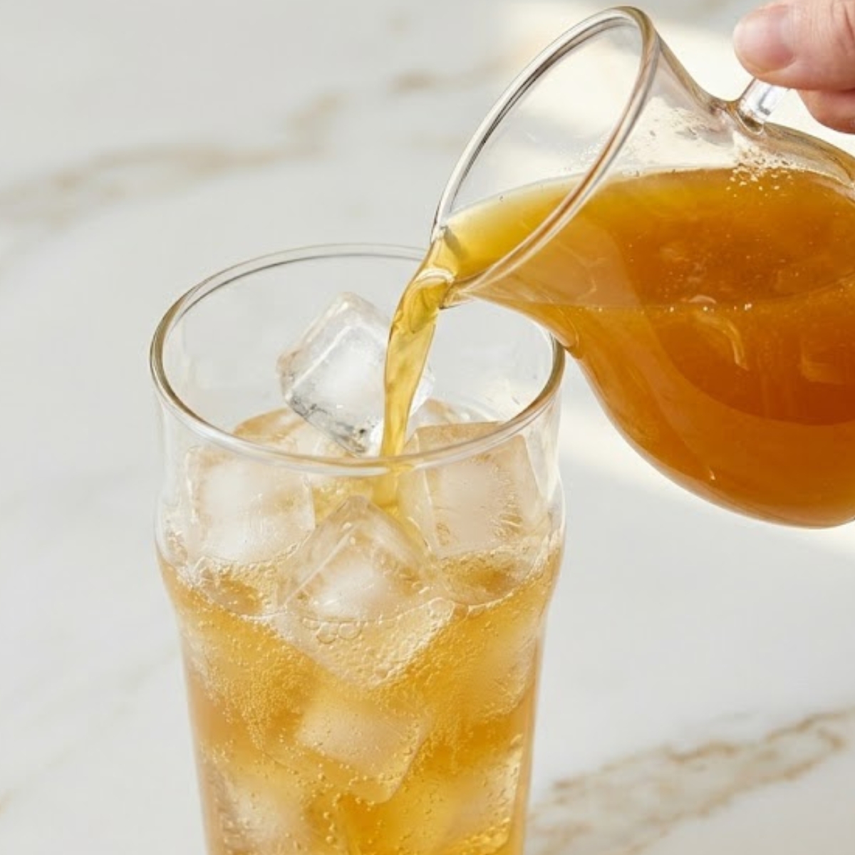A close-up view of golden pineapple soda being poured slowly into a tall glass filled with ice cubes, set against the indistinct gold-veined marble background.