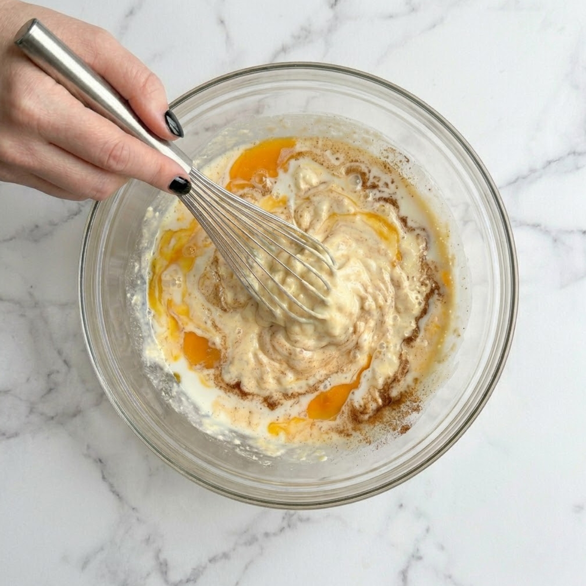 Extreme close up of hands with black nails whisking egg and cottage cheese custard in a glass bowl.