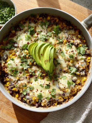 A skillet of Salsa Verde Chicken & Rice with corn, black beans, and melted cheese, topped with sliced avocado, fresh cilantro, and red pepper flakes, served on a wooden board.
