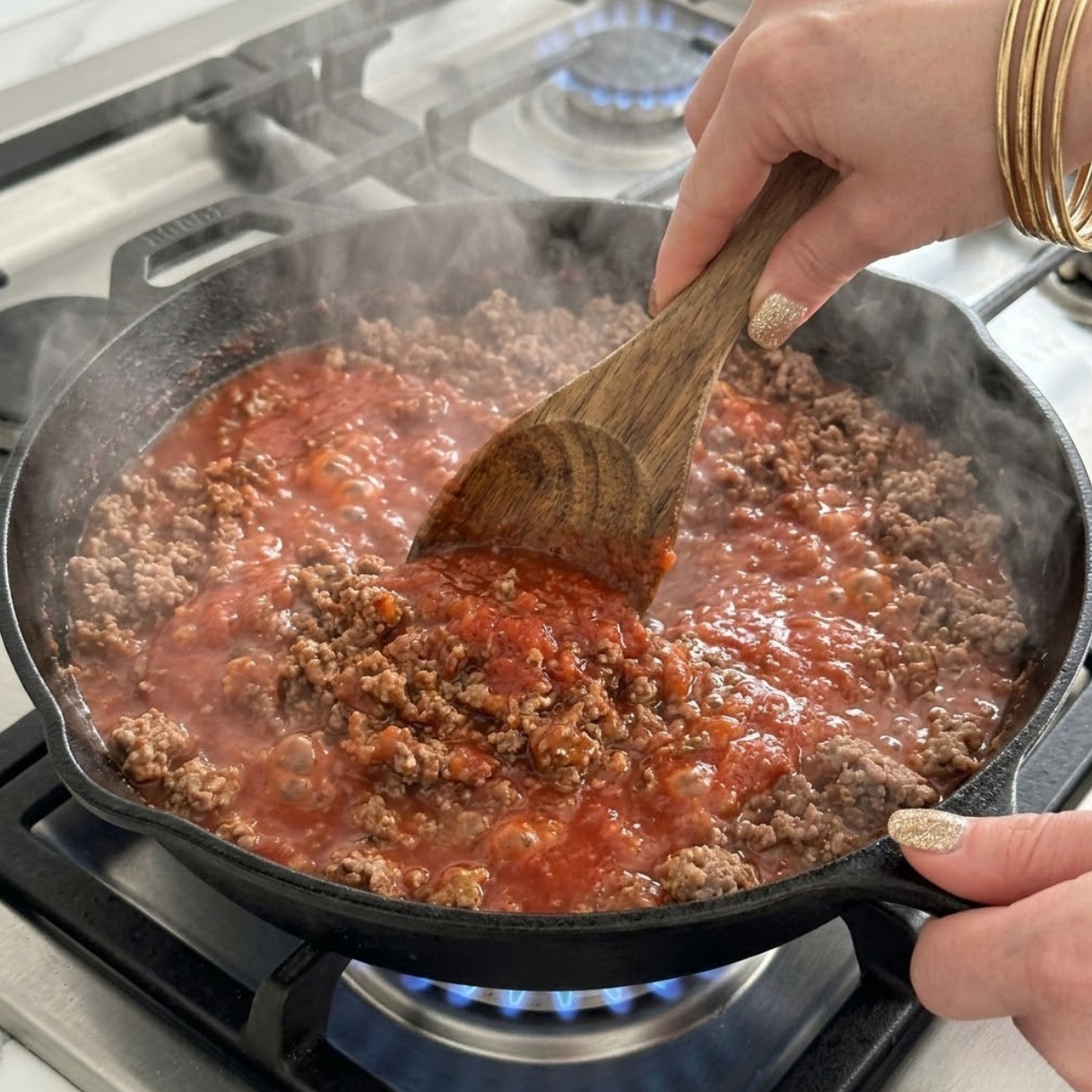 Tight zoom of hands with gold nails stirring bubbling marinara sauce into browned ground beef inside a hot skillet.