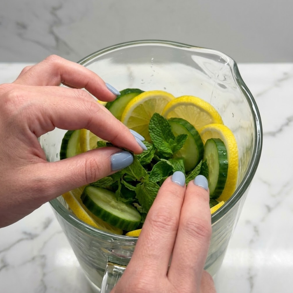 Close up view of a woman's hands with fog blue nails using a sharp knife to slice an English cucumber into thick rounds on a cutting board over a marble counter.