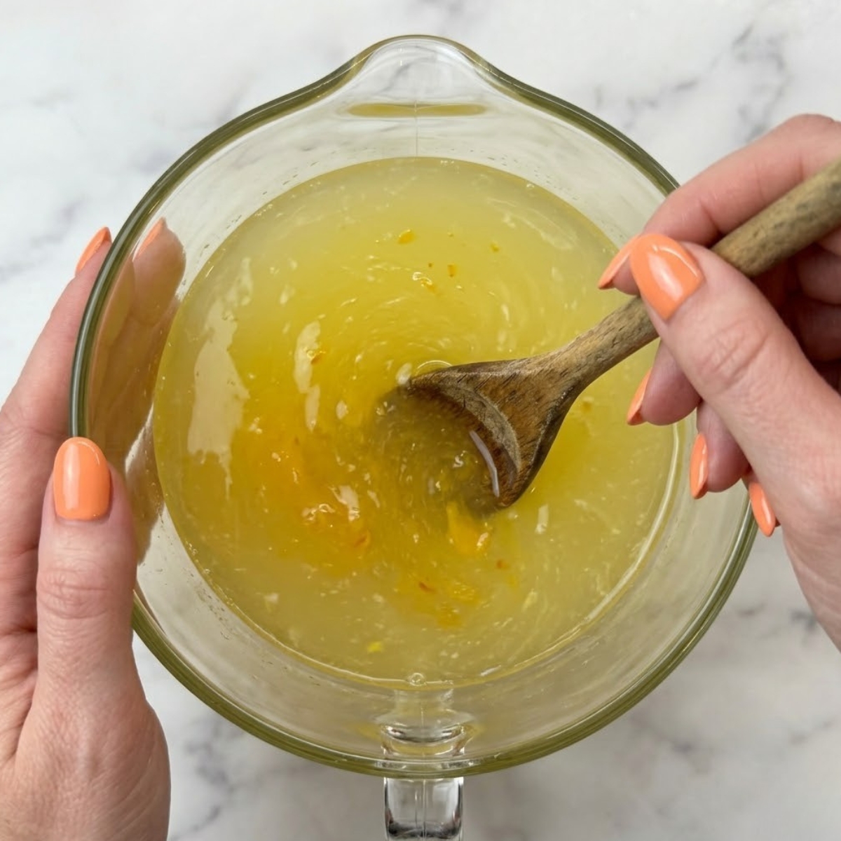 Tight zoom of hands with orange nails using a long wooden spoon to mix fresh yellow peach lemonade inside a large glass pitcher.