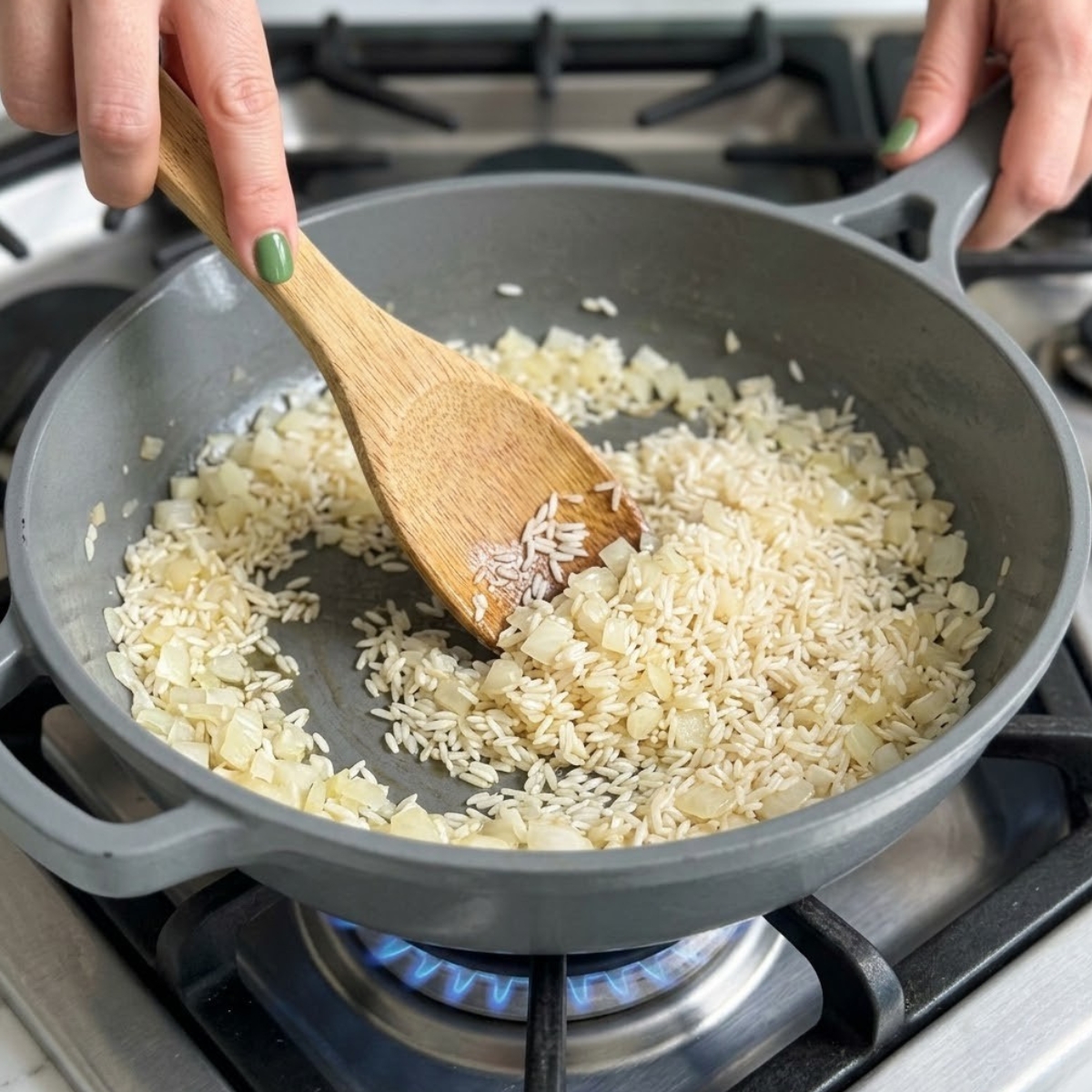 Extreme close-up of hands with green nails using a wooden spoon to sauté white rice and onions in a large skillet over a gas flame.