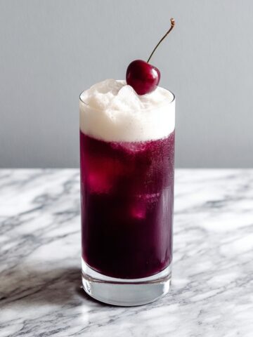 A tall clear highball glass filled with a deep burgundy cherry rum cocktail over ice, topped with a thick billowing white coconut foam layer and garnished with a single fresh dark cherry on a stem, placed on a white and gray veined marble surface against a clean light gray background.