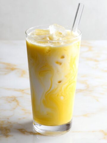 A tall clear highball glass filled with a vibrant golden yellow tropical dirty soda, showcasing beautiful swirling ribbons of cream blending into the bright mango or pineapple base, packed with ice cubes and a clear glass straw, set on a white and gold veined marble surface against a light gray background.