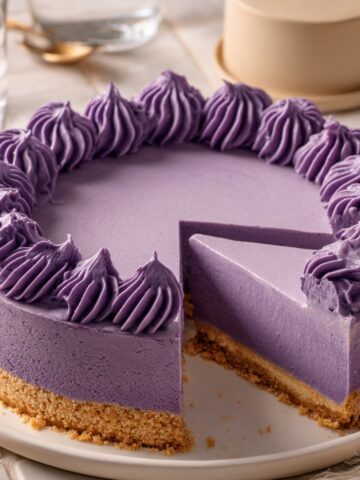 A sliced Ube Cheesecake with a smooth purple ube filling, graham cracker crust, and decorative purple ube cream piped around the edges, served on a white plate.