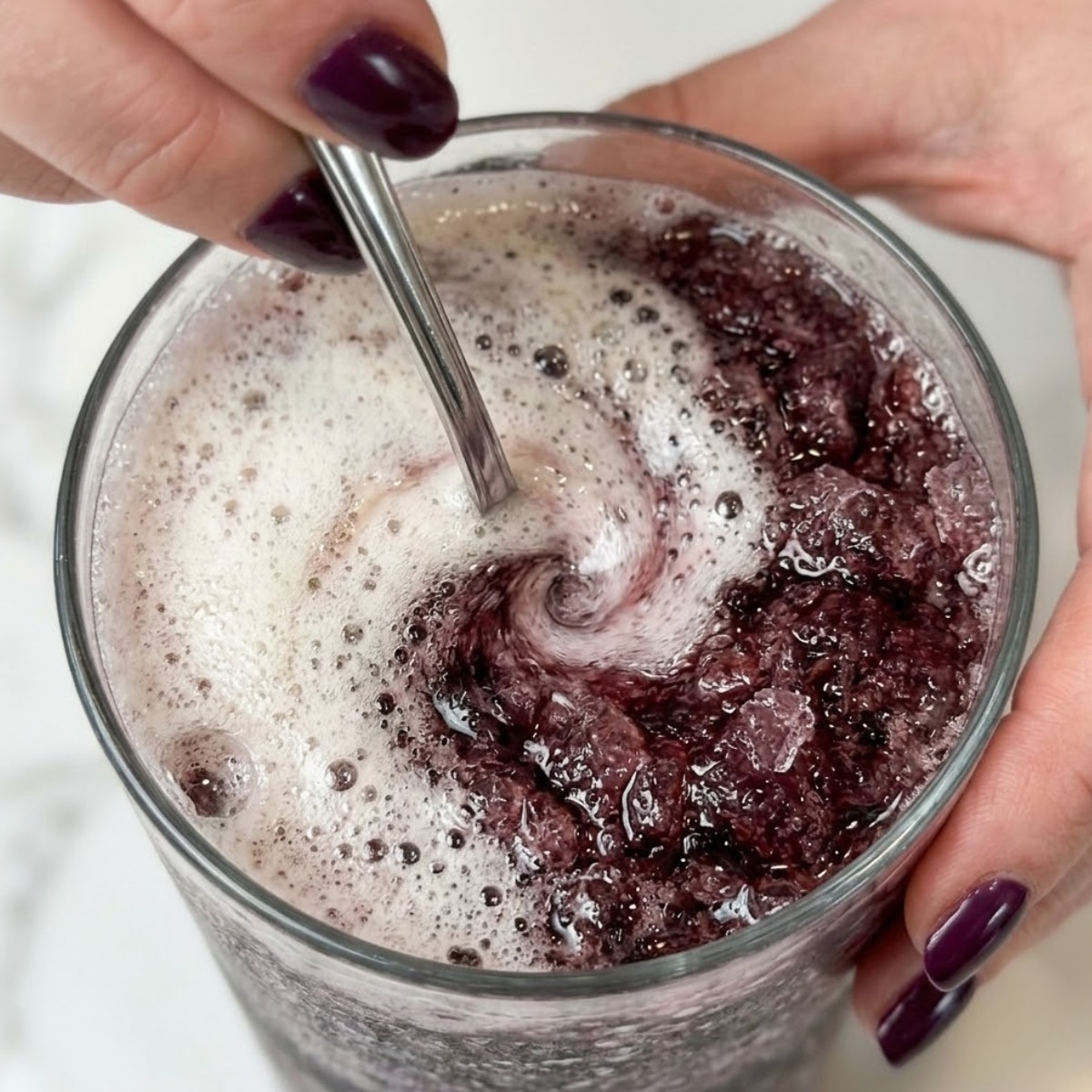 Extreme close-up of hands with plum nails stirring a fizzy purple cherry slushie with a silver spoon.