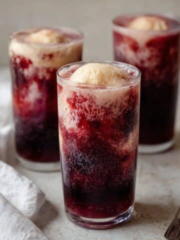 Three tall clear glasses filled with deep crimson cherry cola slushies topped with a round scoop of creamy vanilla ice cream creating a fizzing float effect, arranged on a light stone surface with a white linen napkin and a silver spoon alongside, against a soft neutral background.