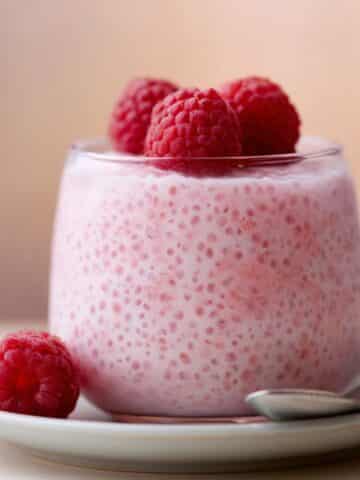 A clear glass filled with delicate pale pink vanilla raspberry chia pudding with visible swollen chia seeds throughout, topped with three plump fresh raspberries, placed on a white ceramic saucer with additional raspberries and a silver spoon alongside, against a soft warm peach-toned blurred background.You said: Vanilla Raspberry Chia Pudding