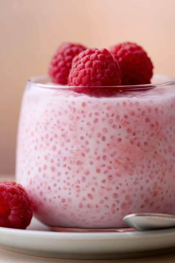 A clear glass filled with delicate pale pink vanilla raspberry chia pudding with visible swollen chia seeds throughout, topped with three plump fresh raspberries, placed on a white ceramic saucer with additional raspberries and a silver spoon alongside, against a soft warm peach-toned blurred background.You said: Vanilla Raspberry Chia Pudding