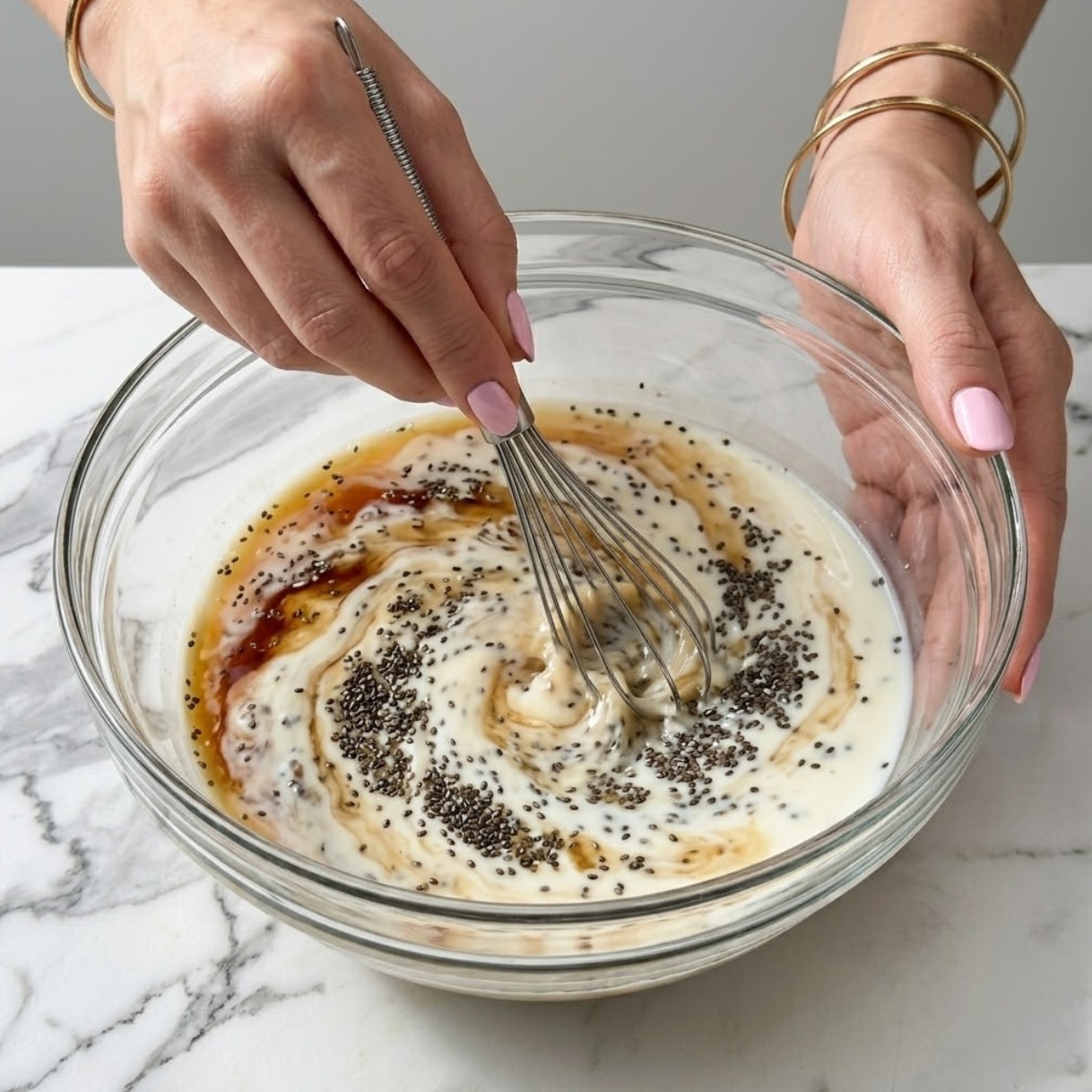Extreme close-up of hands with pink nails whisking black chia seeds into milk inside a glass bowl.