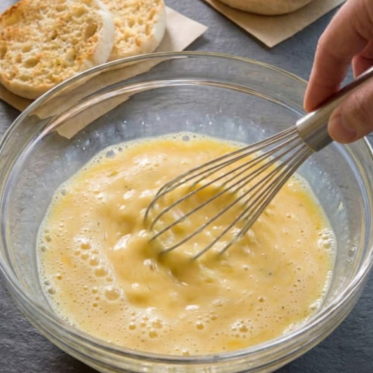 A medium close-up view of a hand vigorously whisking the raw eggs, milk, garlic powder, and black pepper (from image_12.png) inside a large glass bowl until frothy, with the background showing the split English muffin halves on parchment squares on the dark slate, matching the specific plating preparation seen in image_11.png.