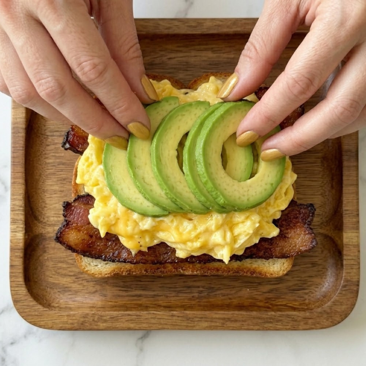 Tight close-up zoom of hands with gold nails layering sliced avocado onto fluffy scrambled eggs and toasted white bread.