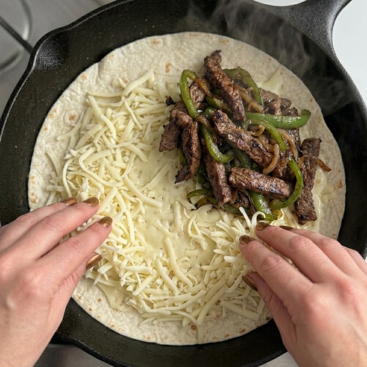Tight close-up zoom of hands with bronze nails layering shredded provolone cheese and charred steak filling onto a flour tortilla inside a pan.