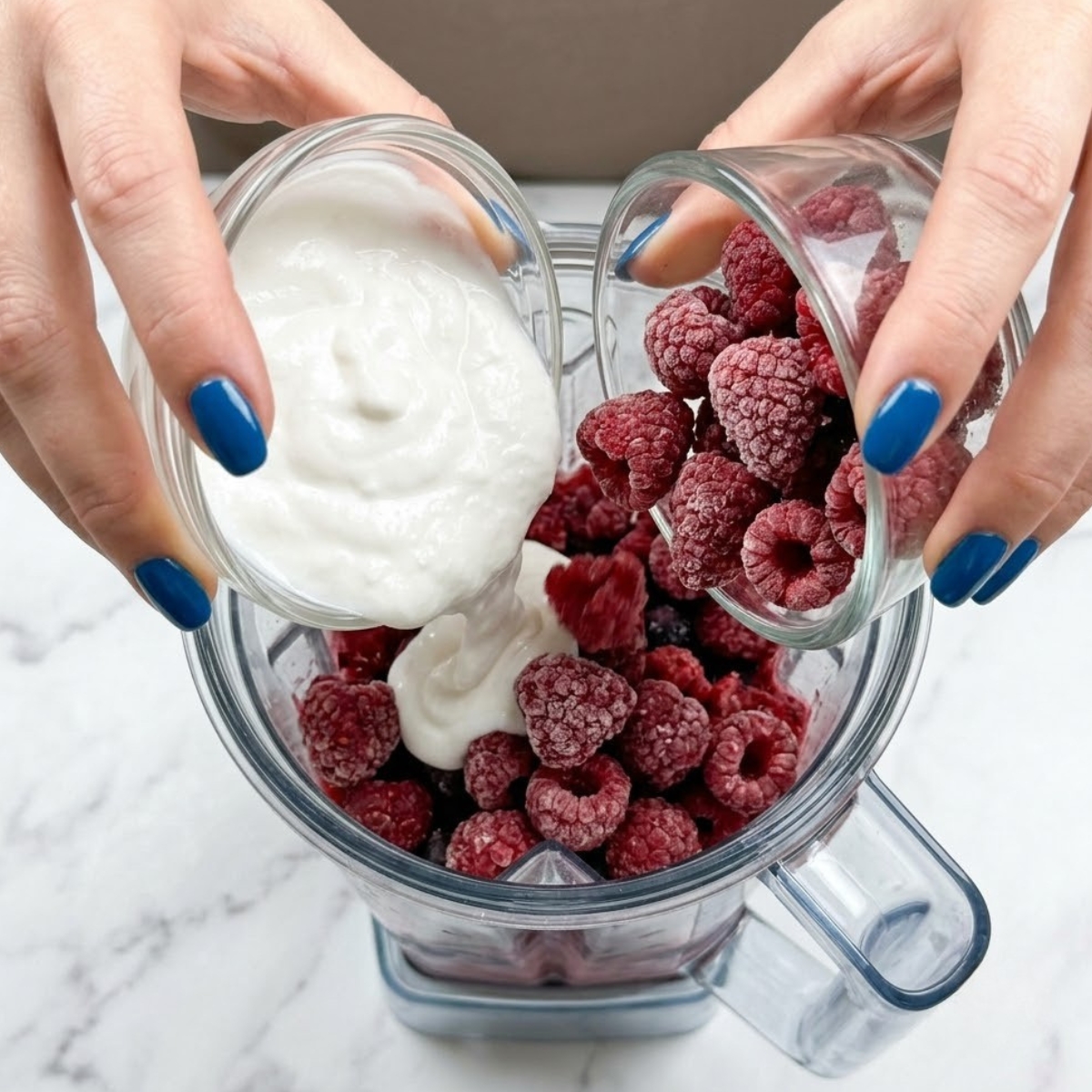 Extreme close-up view of hands with blue nails adding frozen red raspberries and coconut cream into a blender jar.