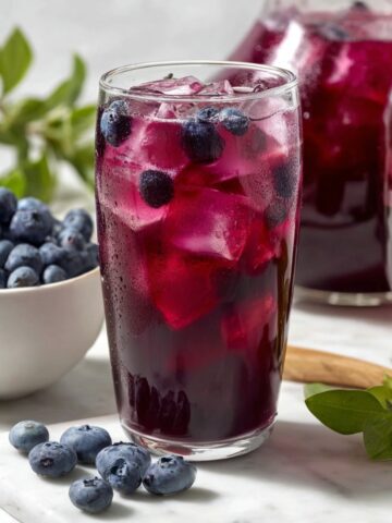 A tall glass of deep purple blueberry iced tea with large ice cubes and fresh blueberries, served alongside a bowl of blueberries, a pitcher of blueberry iced tea, and fresh herb sprigs on a marble surface.