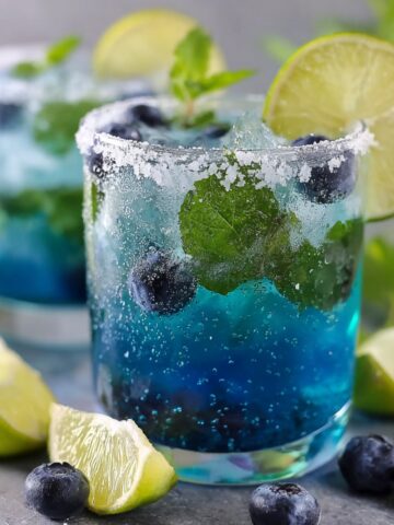 Two sparkling blue blueberry infused water glasses with a salted rim, fresh mint leaves, and whole blueberries, garnished with a lime wheel, with lime wedges and scattered blueberries on a gray surface.