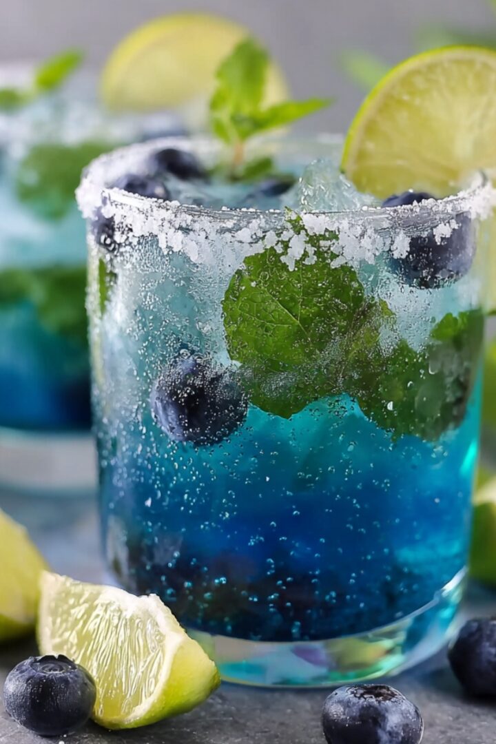 Two sparkling blue blueberry infused water glasses with a salted rim, fresh mint leaves, and whole blueberries, garnished with a lime wheel, with lime wedges and scattered blueberries on a gray surface.
