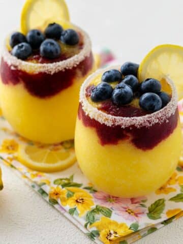 Two sugar-rimmed glasses filled with layered yellow lemon and deep purple blueberry slushies, topped with fresh blueberries and a lemon slice, set on a floral napkin surrounded by fresh lemons.