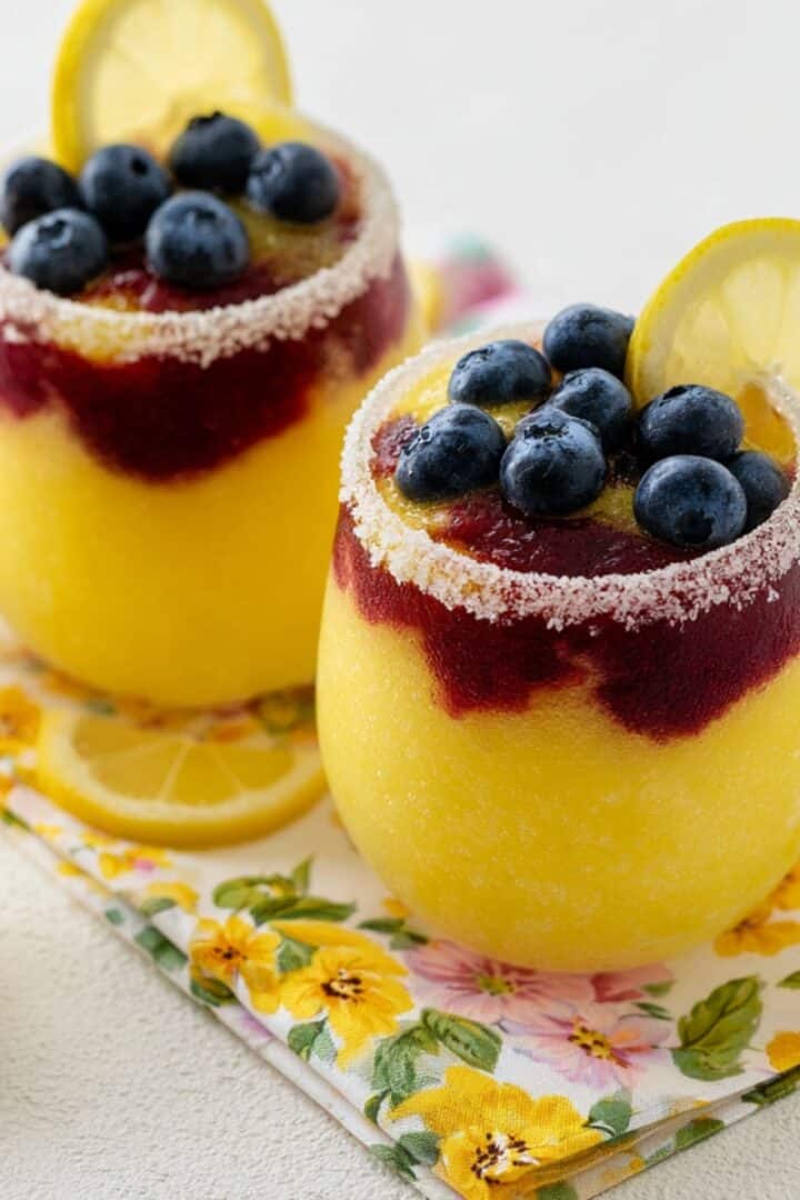 Two sugar-rimmed glasses filled with layered yellow lemon and deep purple blueberry slushies, topped with fresh blueberries and a lemon slice, set on a floral napkin surrounded by fresh lemons.