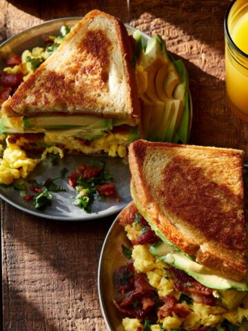 Two plates of breakfast club sandwiches on toasted bread filled with scrambled eggs, crispy bacon, and avocado slices, served with a glass of fresh orange juice on a rustic wooden table.