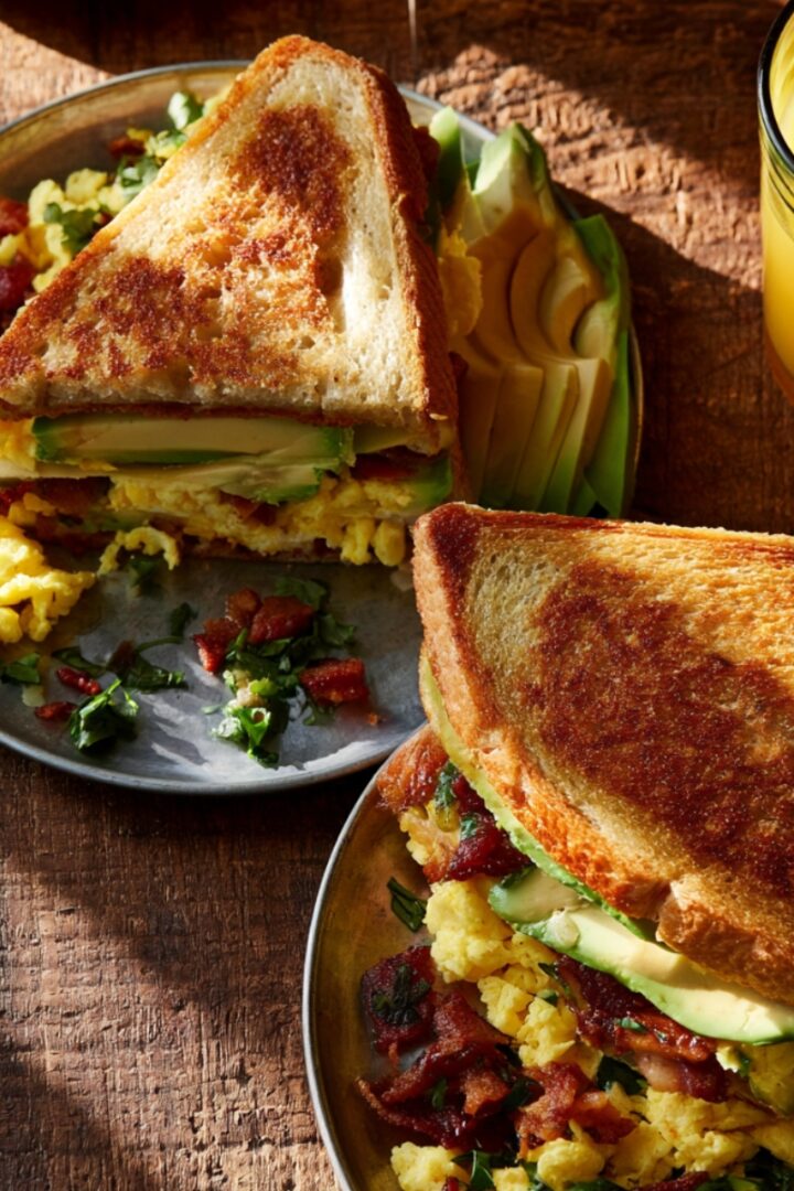 Two plates of breakfast club sandwiches on toasted bread filled with scrambled eggs, crispy bacon, and avocado slices, served with a glass of fresh orange juice on a rustic wooden table.
