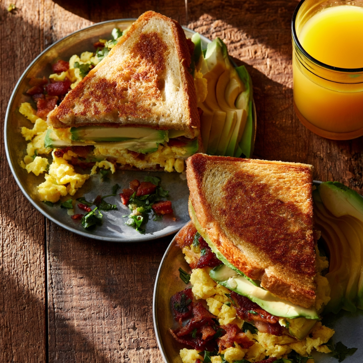 Two plates of breakfast club sandwiches on toasted bread filled with scrambled eggs, crispy bacon, and avocado slices, served with a glass of fresh orange juice on a rustic wooden table.