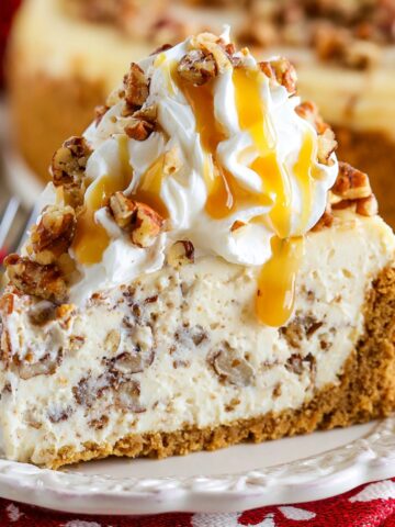 A slice of browned butter pecan cheesecake on a white plate, with a thick graham cracker crust, creamy pecan-studded filling, and topped with whipped cream, chopped toasted pecans, and a drizzle of caramel sauce.