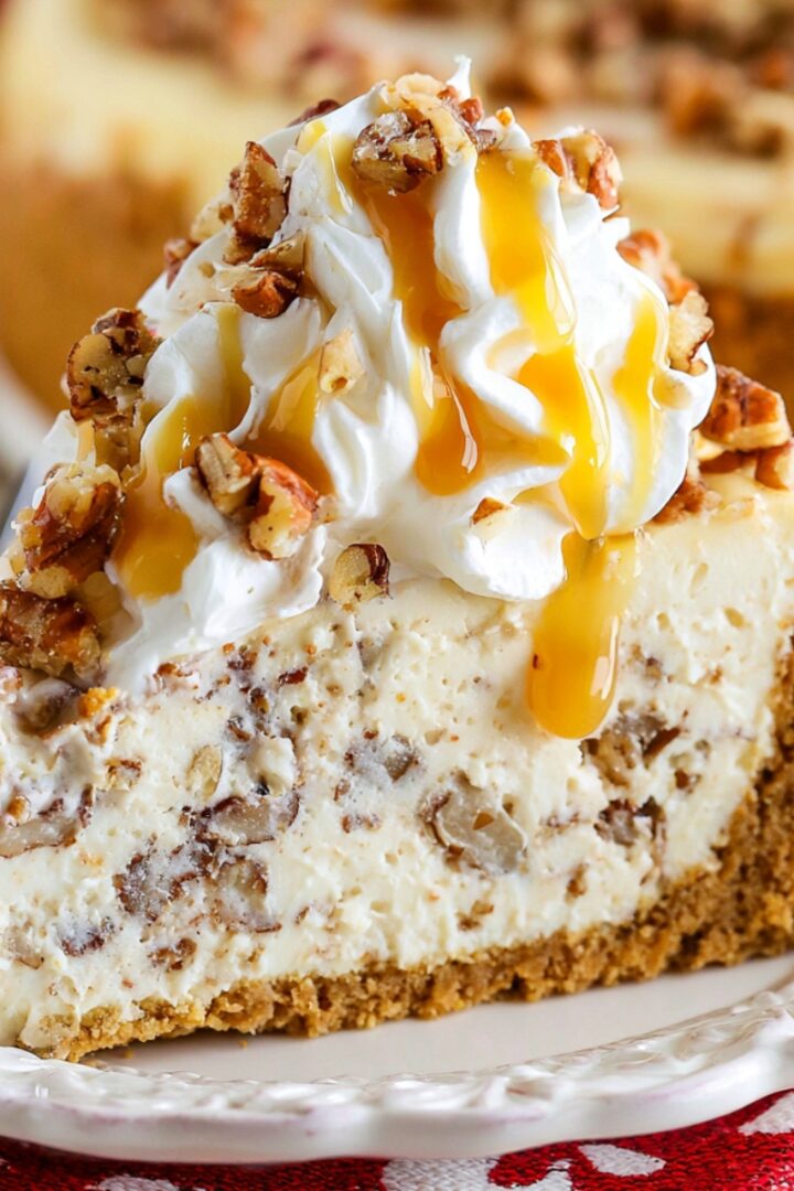 A slice of browned butter pecan cheesecake on a white plate, with a thick graham cracker crust, creamy pecan-studded filling, and topped with whipped cream, chopped toasted pecans, and a drizzle of caramel sauce.