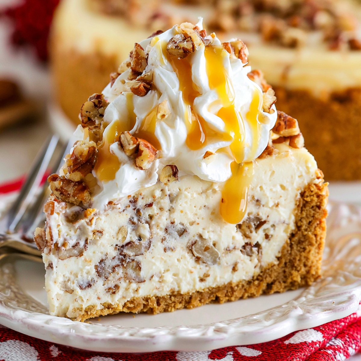 A slice of browned butter pecan cheesecake on a white plate, with a thick graham cracker crust, creamy pecan-studded filling, and topped with whipped cream, chopped toasted pecans, and a drizzle of caramel sauce.
