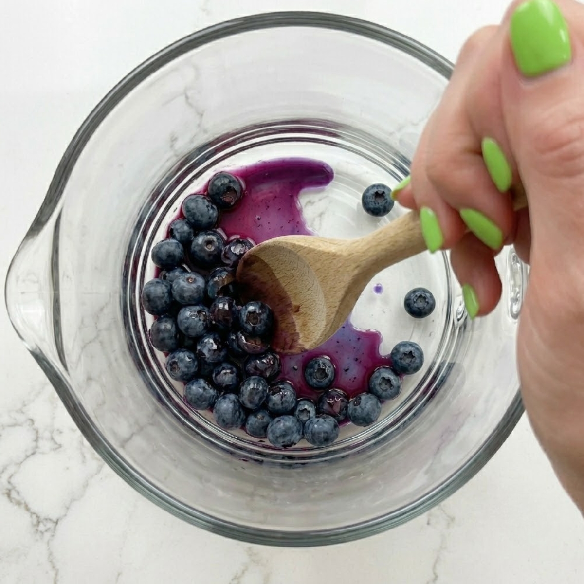 Extreme close-up of hands with green nails using a wooden spoon to gently muddle and bruise fresh blueberries inside a glass pitcher.