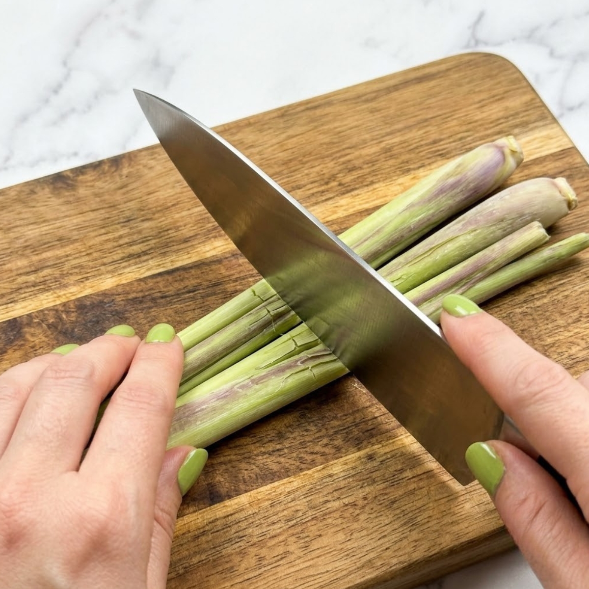 Extreme close-up view of hands with green nails using the flat side of a chef's knife to bruise fresh lemongrass stalks on a cutting board.