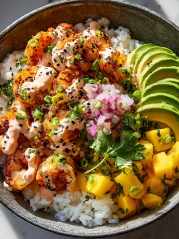 A rustic ceramic bowl with charred seasoned shrimp and steamed white rice, topped with sliced avocado, diced mango, pickled red onion, sliced green onions, cilantro, black sesame seeds, and a drizzle of spicy pink sauce.