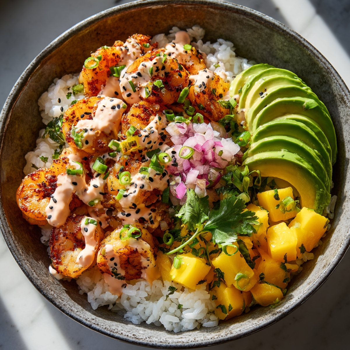 A rustic ceramic bowl with charred seasoned shrimp and steamed white rice, topped with sliced avocado, diced mango, pickled red onion, sliced green onions, cilantro, black sesame seeds, and a drizzle of spicy pink sauce.