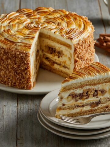 A tall Cinnabon cinnamon swirl cheesecake with a cinnamon crumb coating, layers of creamy cheesecake and cinnamon swirl filling, topped with toasted swirled meringue, with a slice plated on white dishes alongside cinnamon sticks on a rustic wooden surface.